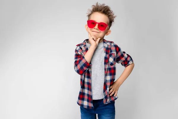 Een knappe jongen in een plaid shirt, grijs shirt en spijkerbroek staat op een grijze achtergrond. Een jongen in rode zonnebril. De jongen gestut zijn kin met twee vingers — Stockfoto