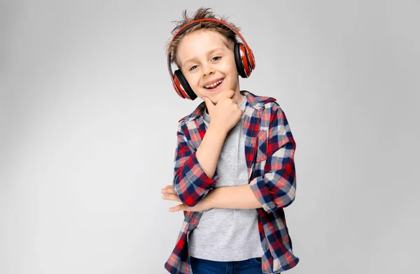 Ένα όμορφο αγόρι σε ένα καρό πουκάμισο, γκρι πουκάμισο και τζιν στέκεται σε γκρι φόντο. Ένα αγόρι στο κόκκινο ακουστικά. Το αγόρι κρατά ένα χέρι στο πηγούνι του. — Φωτογραφία Αρχείου