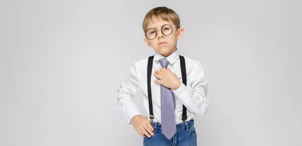 Un niño encantador con una camisa blanca, tirantes, corbata y pantalones vaqueros ligeros se levanta sobre un fondo gris. Un niño sonríe y sostiene su corbata de mano — Foto de Stock