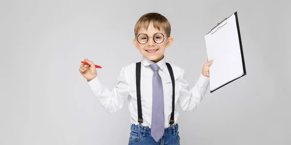 Un niño encantador con una camisa blanca, tirantes, corbata y pantalones vaqueros ligeros se levanta sobre un fondo gris. El chico sostiene un bolígrafo y sábanas para notas — Foto de Stock