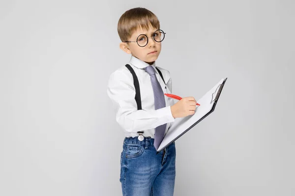 Un niño encantador con una camisa blanca, tirantes, corbata y pantalones vaqueros ligeros se levanta sobre un fondo gris. El chico sostiene un bolígrafo y sábanas para notas — Foto de Stock