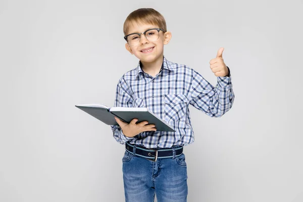 Un niño encantador con una camisa con incrustaciones y pantalones vaqueros ligeros se levanta sobre un fondo gris. El chico tiene un libro en sus manos. Niño con gafas — Foto de Stock