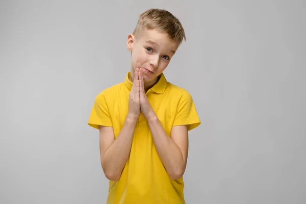 Portret van schattige kleine blonde Kaukasische jongen in geel t-shirt hoop vragen om vergeving op grijze achtergrond — Stockfoto