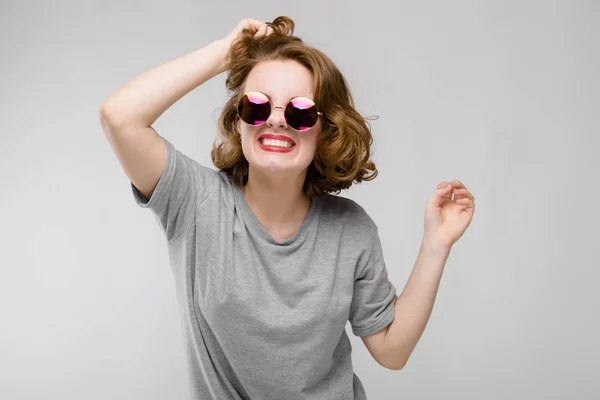 Charmantes junges Mädchen in einem grauen T-Shirt vor grauem Hintergrund. fröhliches Mädchen mit runder Brille hält eine Hand voller Haare — Stockfoto