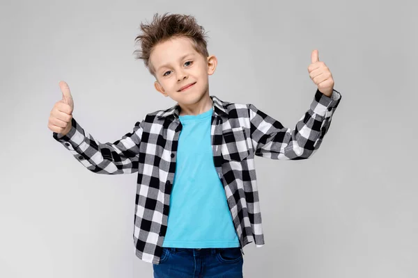 A handsome boy in a plaid shirt, blue shirt and jeans stands on a gray background. The boy smiles and shows class — Stock Photo, Image