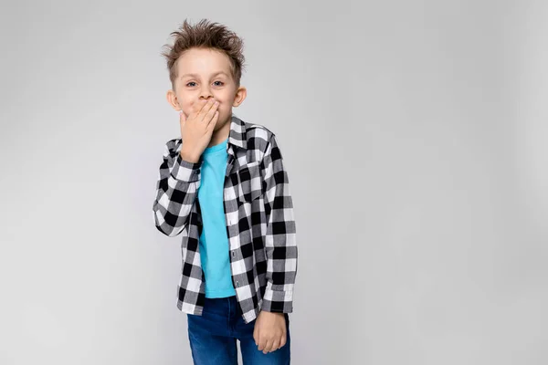 Un chico guapo con camisa a cuadros, camisa azul y pantalones vaqueros está parado sobre un fondo gris. El niño sonríe y se cubre la boca con la mano — Foto de Stock