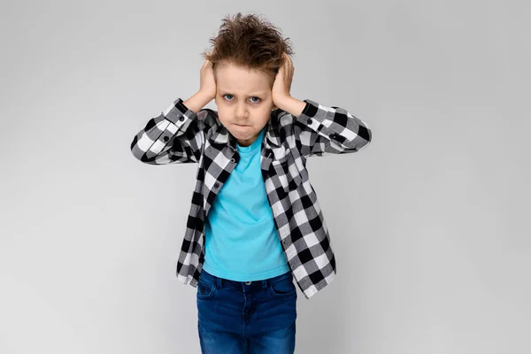 En stilig pojke i en rutig skjorta, ljusblå skjorta och jeans står på en grå bakgrund. Pojken viks armarna över bröstet. Pojken stängt öronen med händerna — Stockfoto