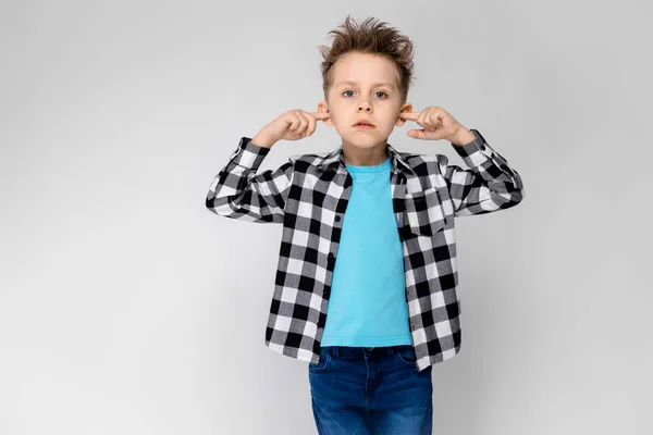 En stilig pojke i en rutig skjorta, ljusblå skjorta och jeans står på en grå bakgrund. Pojken viks armarna över bröstet. Pojken omfattas öronen med fingrarna — Stockfoto