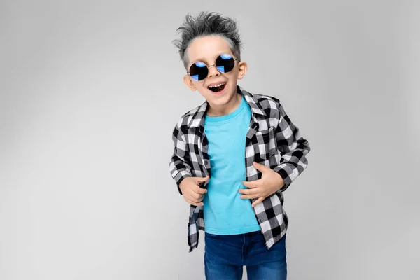 Ένα όμορφο αγόρι σε ένα καρό πουκάμισο, μπλε πουκάμισο και τζιν στέκεται σε γκρι φόντο. Το αγόρι φορώντας στρογγυλά γυαλιά. Το αγόρι χαμογελά. Το αγόρι έχει ένα χρώμα της στάχτης μαλλιά — Φωτογραφία Αρχείου