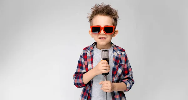 En stilig pojke i en rutig skjorta, grå skjorta och jeans står på en grå bakgrund. En pojke som bär solglasögon. Den rödhåriga pojken lindade tråden runt armen. Pojken håller en mikrofon i handen — Stockfoto