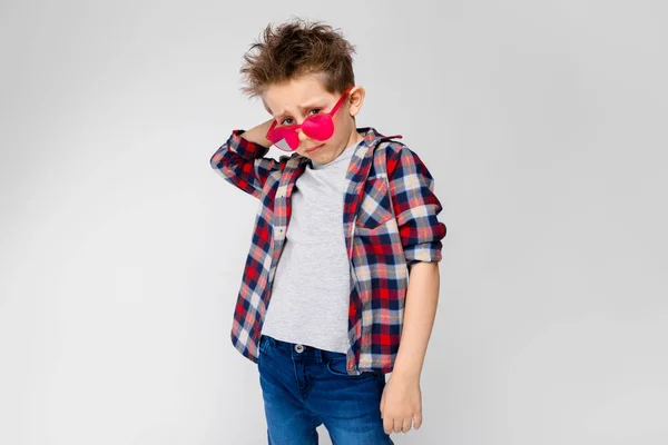 Um menino bonito em uma camisa xadrez, camisa cinza e jeans fica em um fundo cinza. Um rapaz de óculos de sol vermelhos. O menino coça a parte de trás da cabeça — Fotografia de Stock