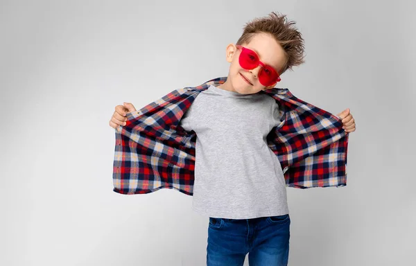 Un chico guapo con una camisa a cuadros, camisa gris y jeans está parado sobre un fondo gris. Un chico con gafas de sol rojas. El chico tira de su camisa hacia atrás . —  Fotos de Stock