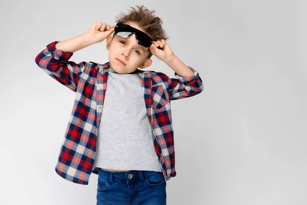 Un chico guapo con una camisa a cuadros, camisa gris y jeans está parado sobre un fondo gris. El chico con gafas de sol negras. El chico sostiene sus gafas. . —  Fotos de Stock