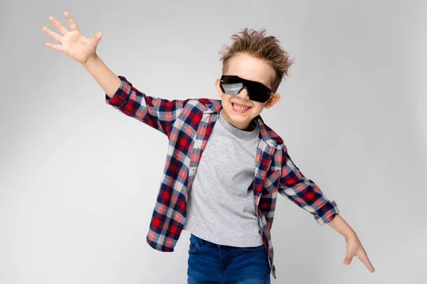 Een knappe jongen in een plaid shirt, grijs shirt en spijkerbroek staat op een grijze achtergrond. De jongen in zwarte zonnebril. De jongen verspreid zijn handen aan de zijkanten. — Stockfoto