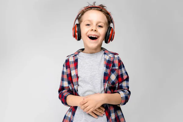 Ένα όμορφο αγόρι σε ένα καρό πουκάμισο, γκρι πουκάμισο και τζιν στέκεται σε γκρι φόντο. Ένα αγόρι στο κόκκινο ακουστικά. Το αγόρι κρατά τα χέρια του στο στομάχι του. Το αγόρι γελάει. — Φωτογραφία Αρχείου