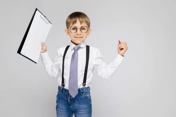 Un niño encantador con una camisa blanca, tirantes, corbata y pantalones vaqueros ligeros se levanta sobre un fondo gris. El chico sostiene un bolígrafo y sábanas para notas — Foto de Stock