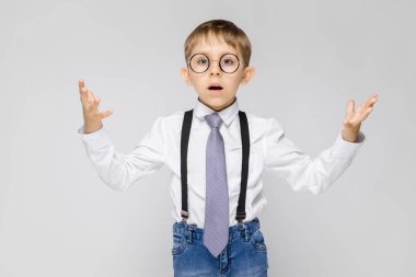 Beyaz bir gömlek, pantolon askısı, kravat ve ışık kot büyüleyici bir çocuk gri bir arka plan üzerinde duruyor. Çocuk elleri her iki yönde yaymak