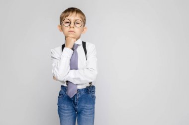 Beyaz bir gömlek, pantolon askısı, kravat ve ışık kot büyüleyici bir çocuk gri bir arka plan üzerinde duruyor. Çocuğun çenesine yumruk üzerinde koymak.