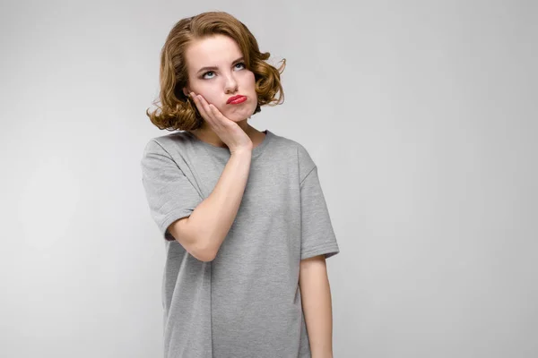 Encantadora joven con una camiseta gris sobre un fondo gris. La chica sujeta su mano a su mejilla — Foto de Stock