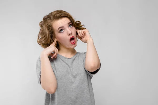 Charming young girl in a gray T-shirt on a gray background. The girl covered her ears with her fingers and stuck out her tongue. — Stock Photo, Image