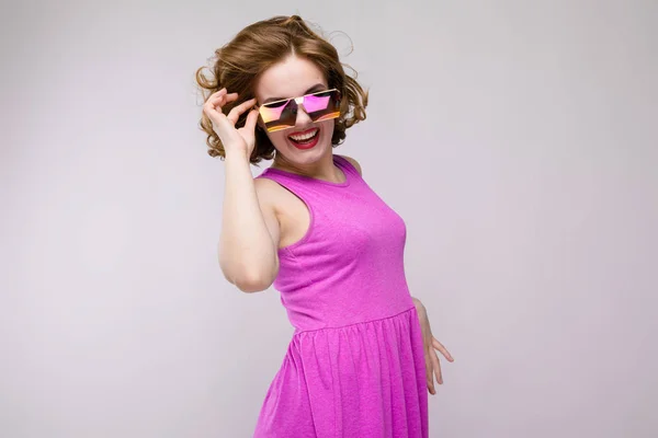 Charmante junge Mädchen in rosa Kleid auf grauem Hintergrund. fröhliches Mädchen mit quadratischen Gläsern. das Mädchen justiert die Brille — Stockfoto