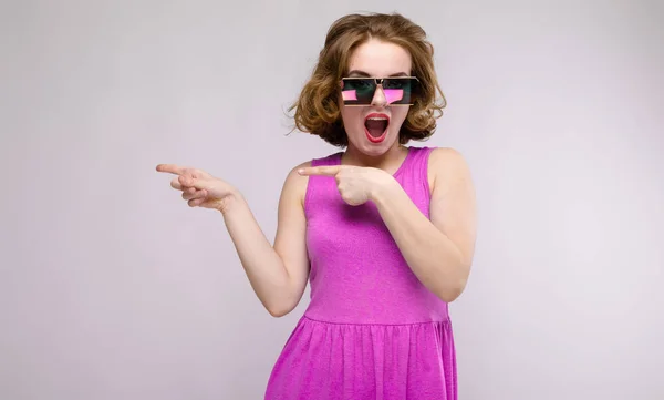 Charmante junge Mädchen in rosa Kleid auf grauem Hintergrund. fröhliches Mädchen mit quadratischen Gläsern. das Mädchen zeigt mit den Fingern zur Seite — Stockfoto
