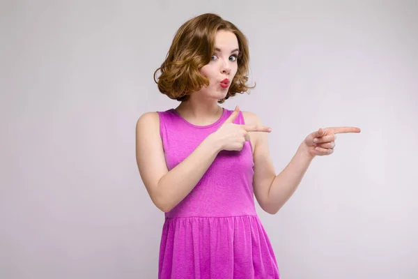 Charmante junge Mädchen in rosa Kleid auf grauem Hintergrund. ein junges Mädchen zeigt ihre Finger zur Seite — Stockfoto