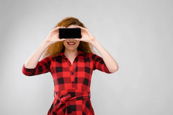 Menina ruiva jovem em uma camisa quadriculada vermelha. Uma jovem cobriu os olhos com um telefone — Fotografia de Stock