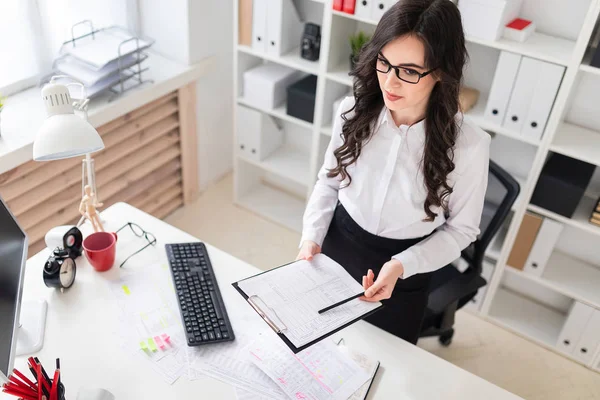 Een mooi jong meisje staat in de buurt van een bureau en houdt in haar handen een blad voor notities en een potlood. — Stockfoto