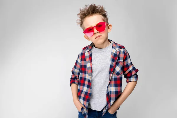 Ένα όμορφο αγόρι σε ένα καρό πουκάμισο, γκρι πουκάμισο και τζιν στέκεται σε γκρι φόντο. Αγόρι σε κόκκινα γυαλιά ηλίου — Φωτογραφία Αρχείου