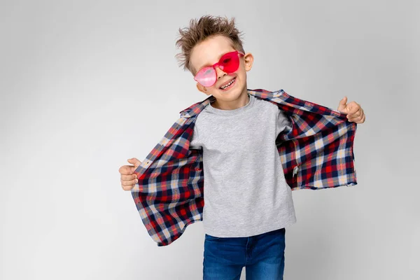 Un chico guapo con una camisa a cuadros, camisa gris y jeans está parado sobre un fondo gris. Un chico con gafas de sol rojas. El chico tira de su camisa hacia atrás . —  Fotos de Stock