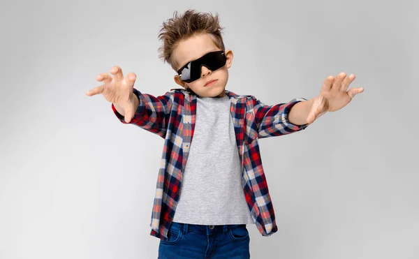 Un chico guapo con una camisa a cuadros, camisa gris y jeans está parado sobre un fondo gris. El chico con gafas de sol negras. El niño extiende los brazos hacia adelante. . — Foto de Stock