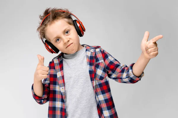 A handsome boy in a plaid shirt, gray shirt and jeans stands on a gray background. A boy in red headphones. The boy points with his fingers to the side. — Stock Photo, Image