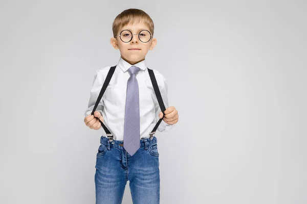 Un niño encantador con una camisa blanca, tirantes, corbata y pantalones vaqueros ligeros se levanta sobre un fondo gris. el chico con gafas tiró de los tirantes — Foto de Stock