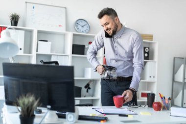 Genç bir adam ofiste masanın, monitör ve dökme kahve fincan içine bakarak onun omuz ile telefon tutarak duruyor.