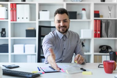 Genç bir adam ofiste bir masada oturur ve elini tutar. Adamın biri tebrik.