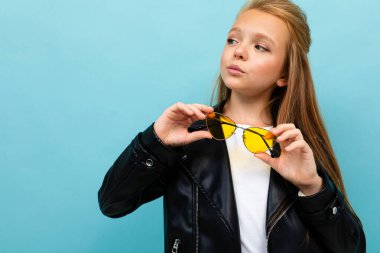 Siyah deri ceketli şirin küçük kız mavi arka planda renkli güneş gözlükleriyle poz veriyor. 