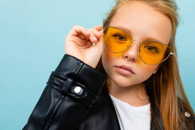 Siyah deri ceketli şirin küçük kız mavi arka planda renkli güneş gözlükleriyle poz veriyor. 