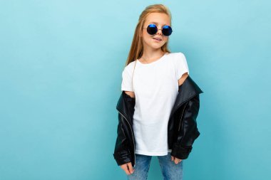 little cool girl in black leather jacket with sunglasses posing against blue background 