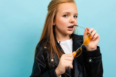 Siyah deri ceketli şirin küçük kız mavi arka planda renkli güneş gözlükleriyle poz veriyor. 