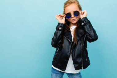 little cool girl in black leather jacket with sunglasses posing against blue background 