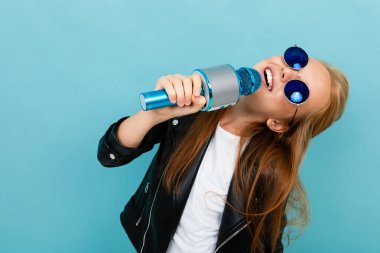 Siyah deri ceketli şirin küçük kız mavi arka planda mikrofonla poz veriyor. 