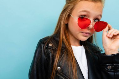 Siyah deri ceketli şirin küçük kız mavi arka planda renkli güneş gözlükleriyle poz veriyor. 