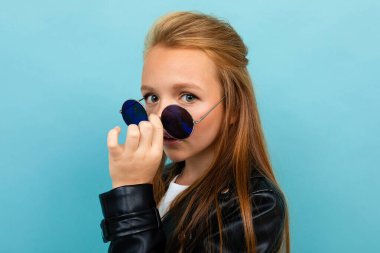 little cool girl in black leather jacket with sunglasses posing against blue background 