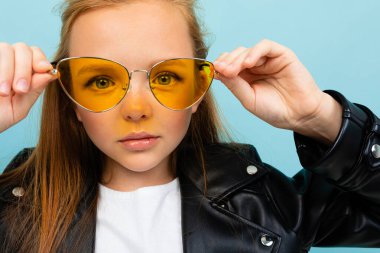 Siyah deri ceketli şirin küçük kız mavi arka planda renkli güneş gözlükleriyle poz veriyor. 