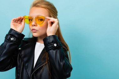 Siyah deri ceketli şirin küçük kız mavi arka planda renkli güneş gözlükleriyle poz veriyor. 