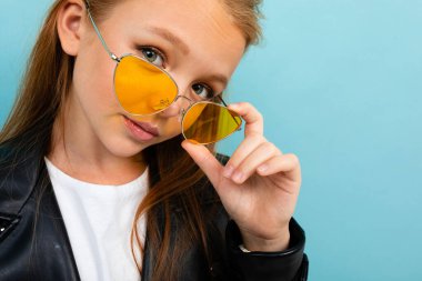Siyah deri ceketli şirin küçük kız mavi arka planda renkli güneş gözlükleriyle poz veriyor. 