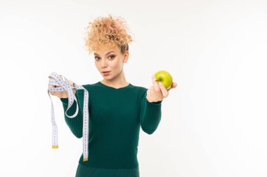 Yeşil elbiseli genç kadın beyaz arka planda elmayla poz veriyor.  