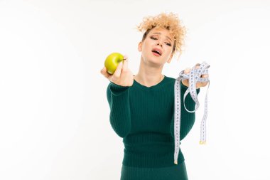 Yeşil elbiseli genç kadın beyaz arka planda elmayla poz veriyor.  