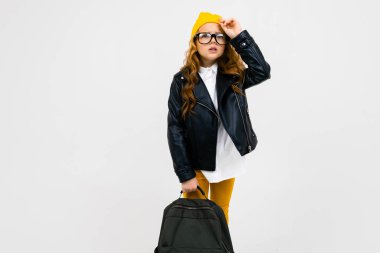 Lazer ceketli havalı küçük kız, arka planda sırt çantasıyla stüdyoda poz veriyor. 